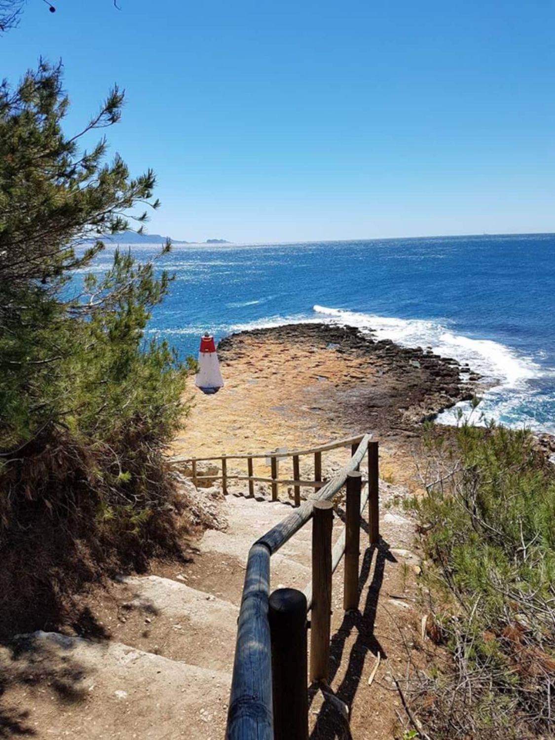 Un escalier en bois mène à une plage bordée d’un rivage rocheux, avec un phare visible au loin.