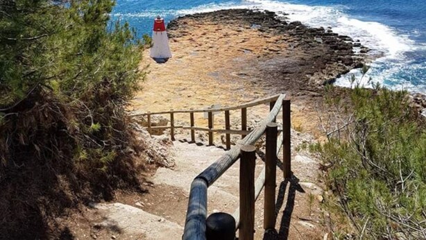 Una escalera de madera que lleva a una playa con costa rocosa y un faro a lo lejos.