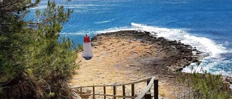 Un escalier en bois mène à une plage bordée d’un rivage rocheux, avec un phare visible au loin.