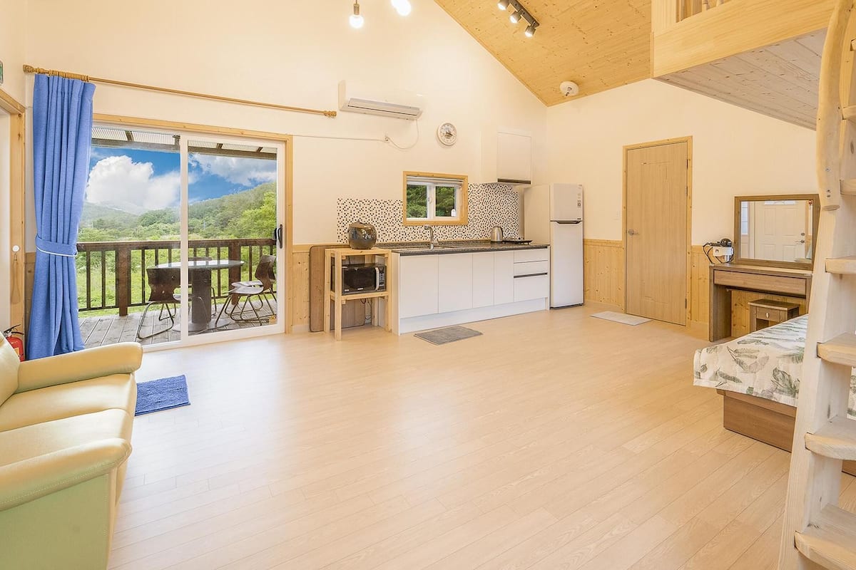 A cozy room with a wooden ceiling, a balcony with a view, a kitchenette, and a bed.