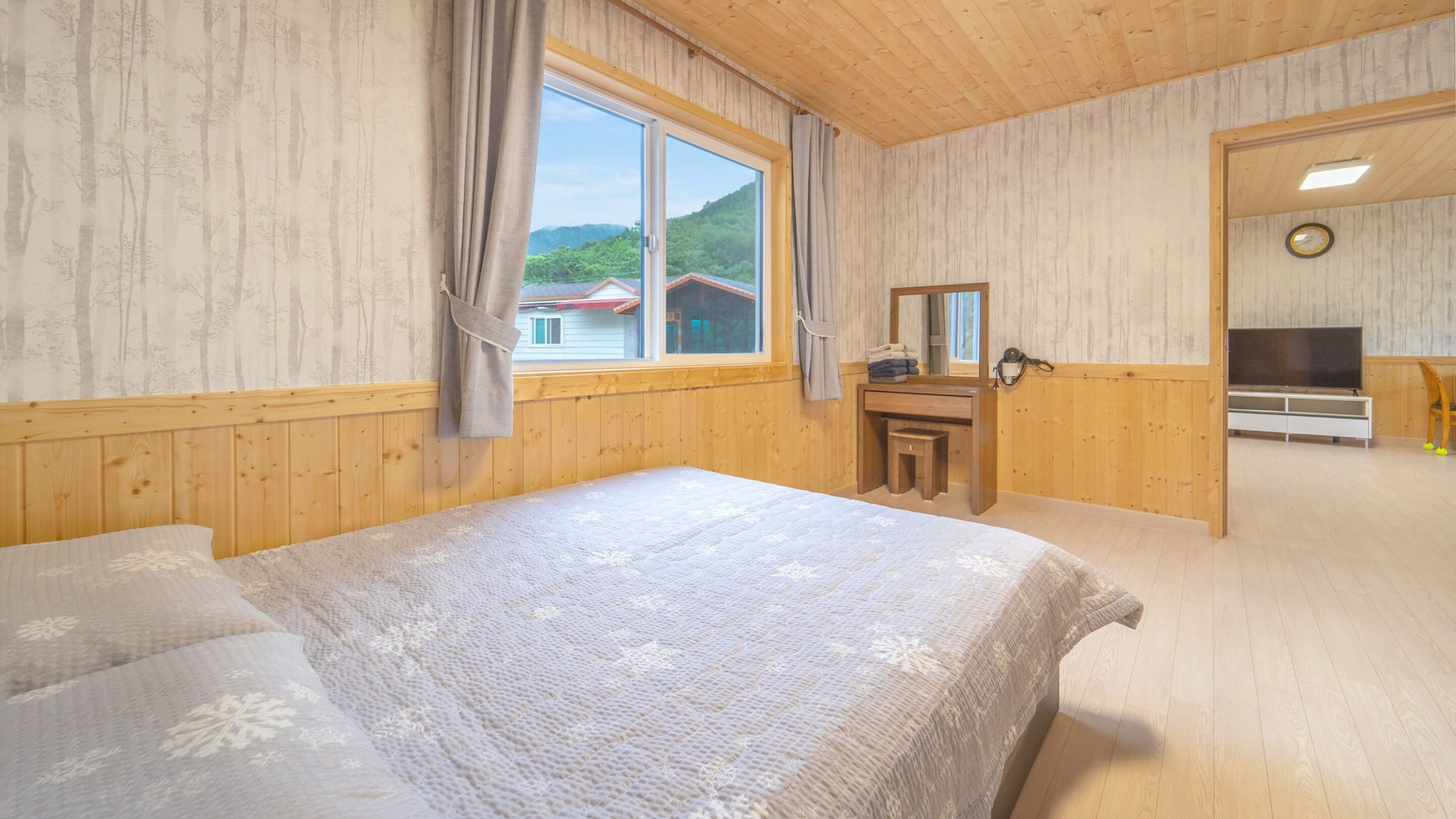 Un dormitorio con una cama grande, un aparador de madera, un espejo, un televisor y una ventana con vistas a casas y árboles.