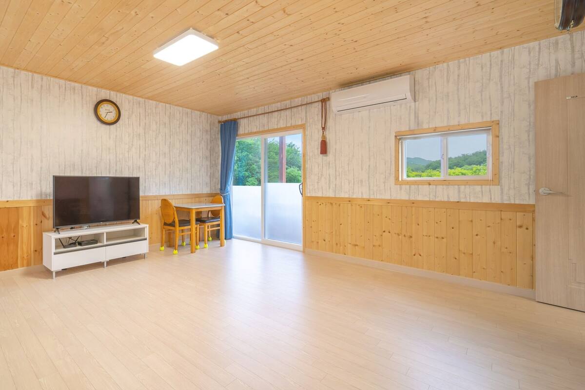 A spacious room with a wooden floor, a flat-screen TV on a white stand, a dining table with chairs, a large window with a view of greenery, and a clock on the wall.