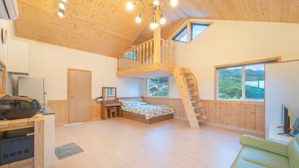 Une pièce spacieuse avec un lit en mezzanine, un escalier en bois et de grandes fenêtres donnant sur l’extérieur.