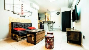 A living room with a black sofa, a wooden chest, and a coffee table. There is a kitchen area with a bar stool and a dining table. A television is mounted on the wall. - INDUSTRIAL#CENTER#STATION#WIFI@SLEEPNTRIPBEZIERS (Béziers)
