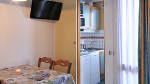 A compact room with a dining table, chairs, a television mounted on the wall, a kitchen area with cabinets and a washing machine, and a window with curtains.
