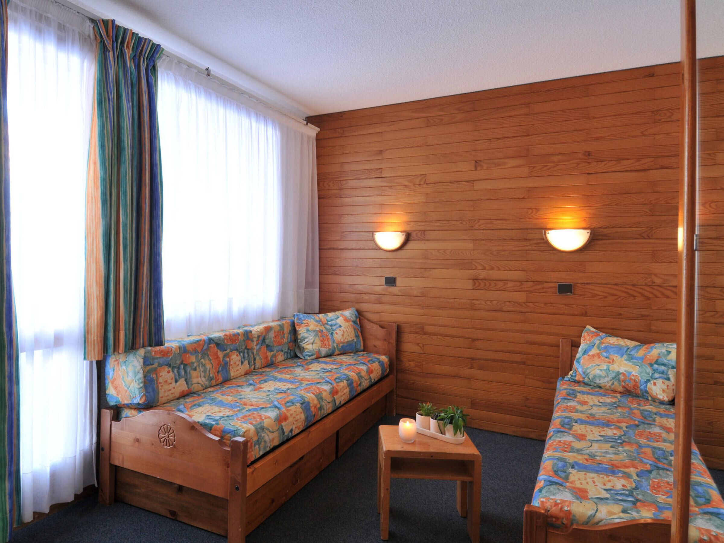 A cozy room with a wooden wall, two sofas with patterned cushions, a small table with a candle and plants, and a window with curtains.