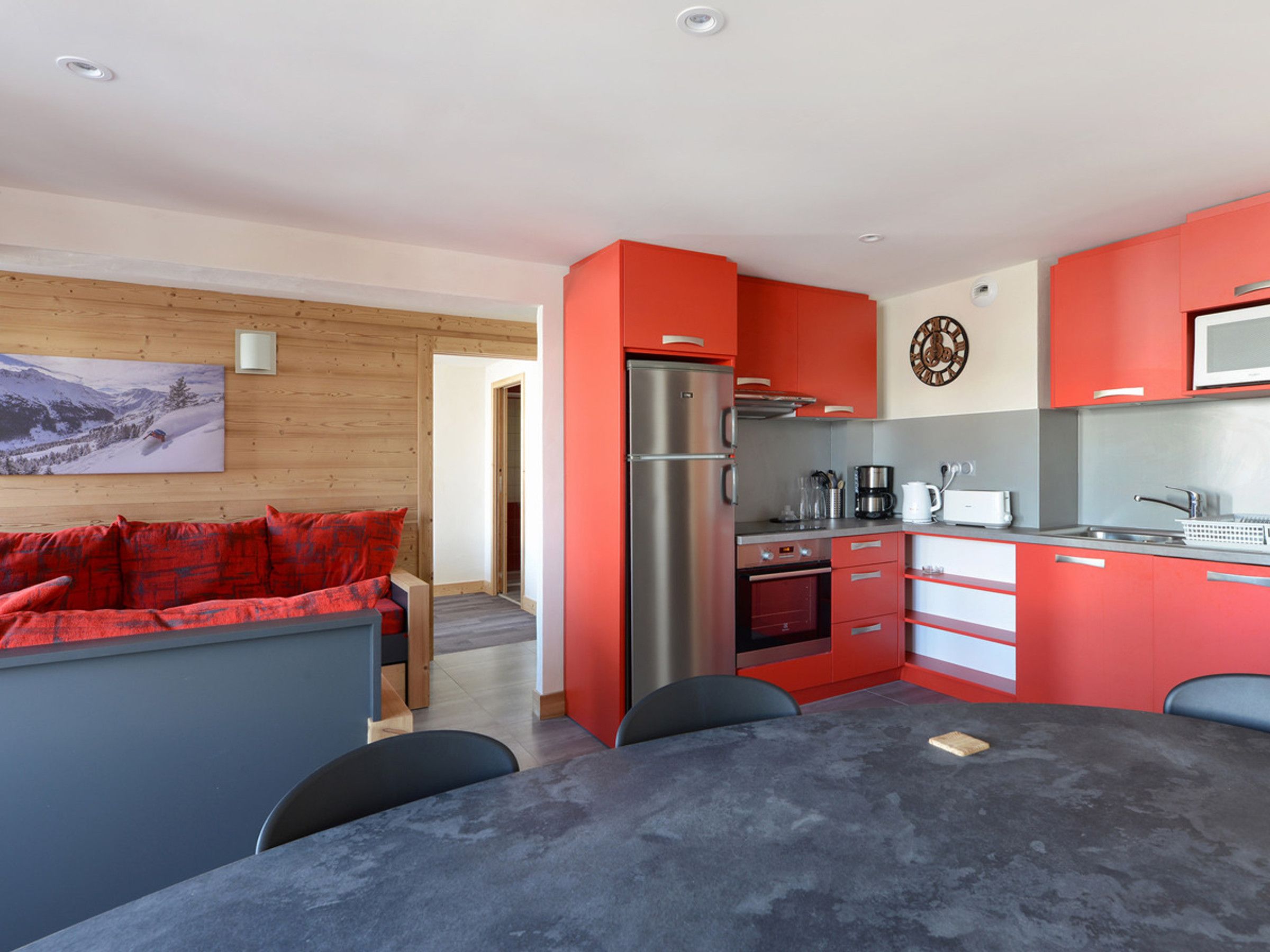 A modern kitchen with red cabinets, stainless steel appliances, and a dining area with a grey table and black chairs.