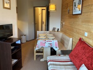 Een gezellige kamer met een televisie, een eettafel gedekt voor twee personen, een houten bankje en een rode kussen.