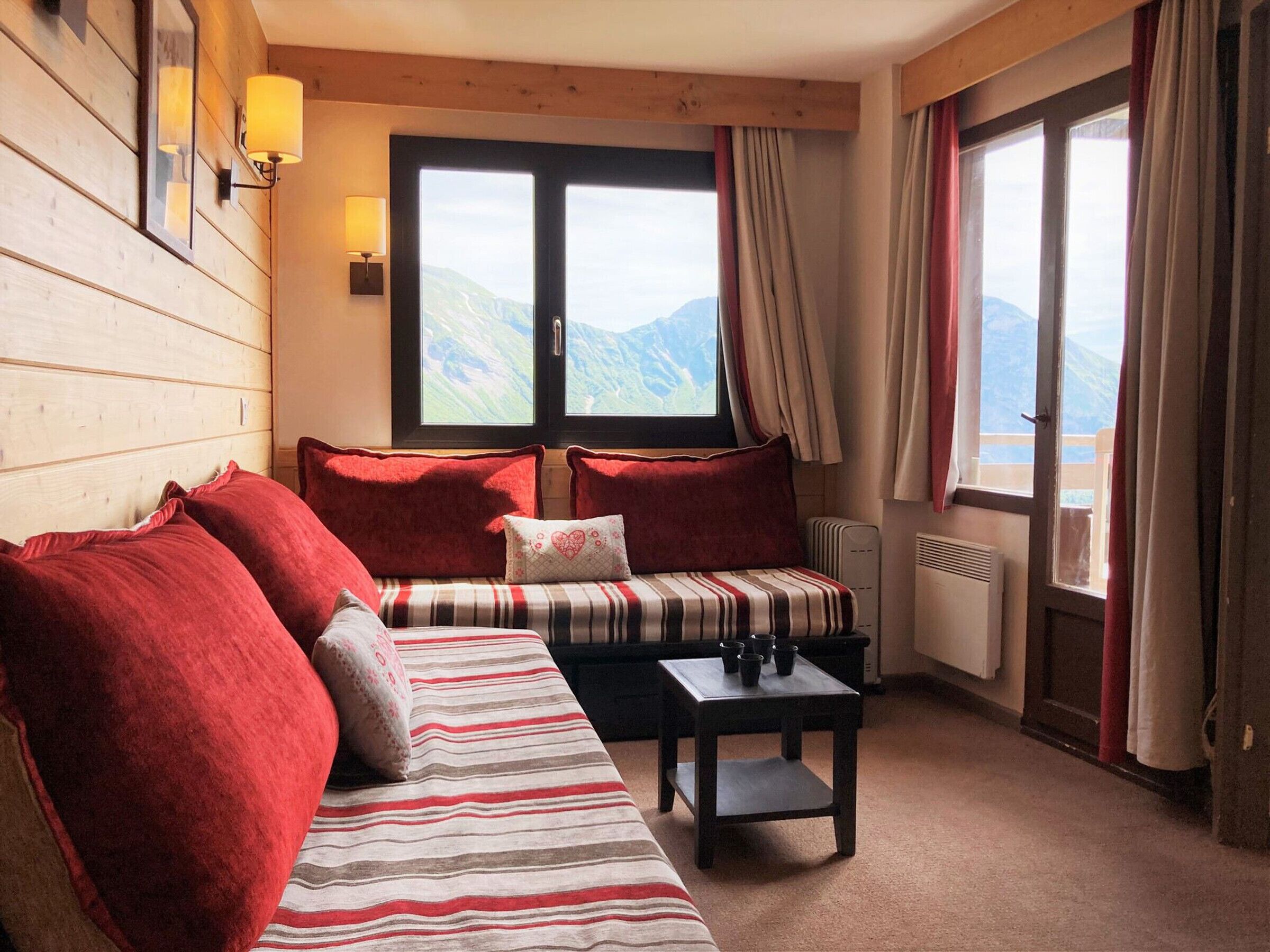 A cozy room with a wooden wall, a striped sofa, red cushions, a small table with cups, and a window with a mountain view.