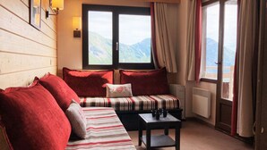Ein gemütliches Zimmer mit einer Holzwand, einem gestreiften Sofa, roten Kissen, einem kleinen Tisch mit Tassen und einem Fenster mit Bergblick.