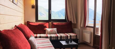 A cozy room with a wooden wall, a striped sofa, red cushions, a small table with cups, and a window with a mountain view.