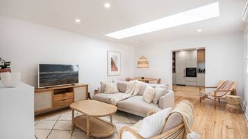 A modern living room with a sofa, coffee table, and TV. There is a dining area with a table and chairs. The kitchen is visible in the background.