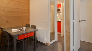 A compact room with a dining table, chairs, a red drawer, and a white door leading to a kitchen with red cabinets.