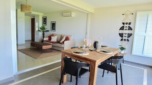 A dining area with a wooden table set for four, surrounded by black chairs. There is a sofa and a coffee table in the background, and a wall-mounted macrame decoration. - Sunset on lagoon from the terrace (Marigot)