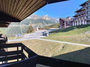 Blick auf Berge von einem Holzbalkon mit Blick auf eine Straße, mit einem Gebäude mit Balkonen und blauem Himmel.