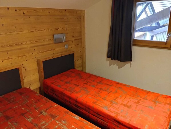 A room with two beds, wooden paneling on the walls, a window with curtains, and a small shelf with a lamp.
