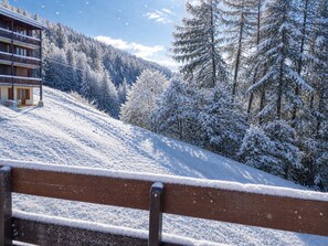 A wooden bench with snow on its backrest, overlooking a snow-covered slope with a building and trees.