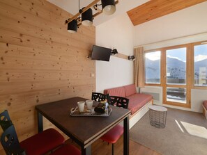 A cozy room with a wooden wall, a TV mounted above a table with red chairs, a sofa with a patterned cover, and a window offering a mountain view. - Renovated Apt 5 pers ski-in/ski-out (Belle Plagne)