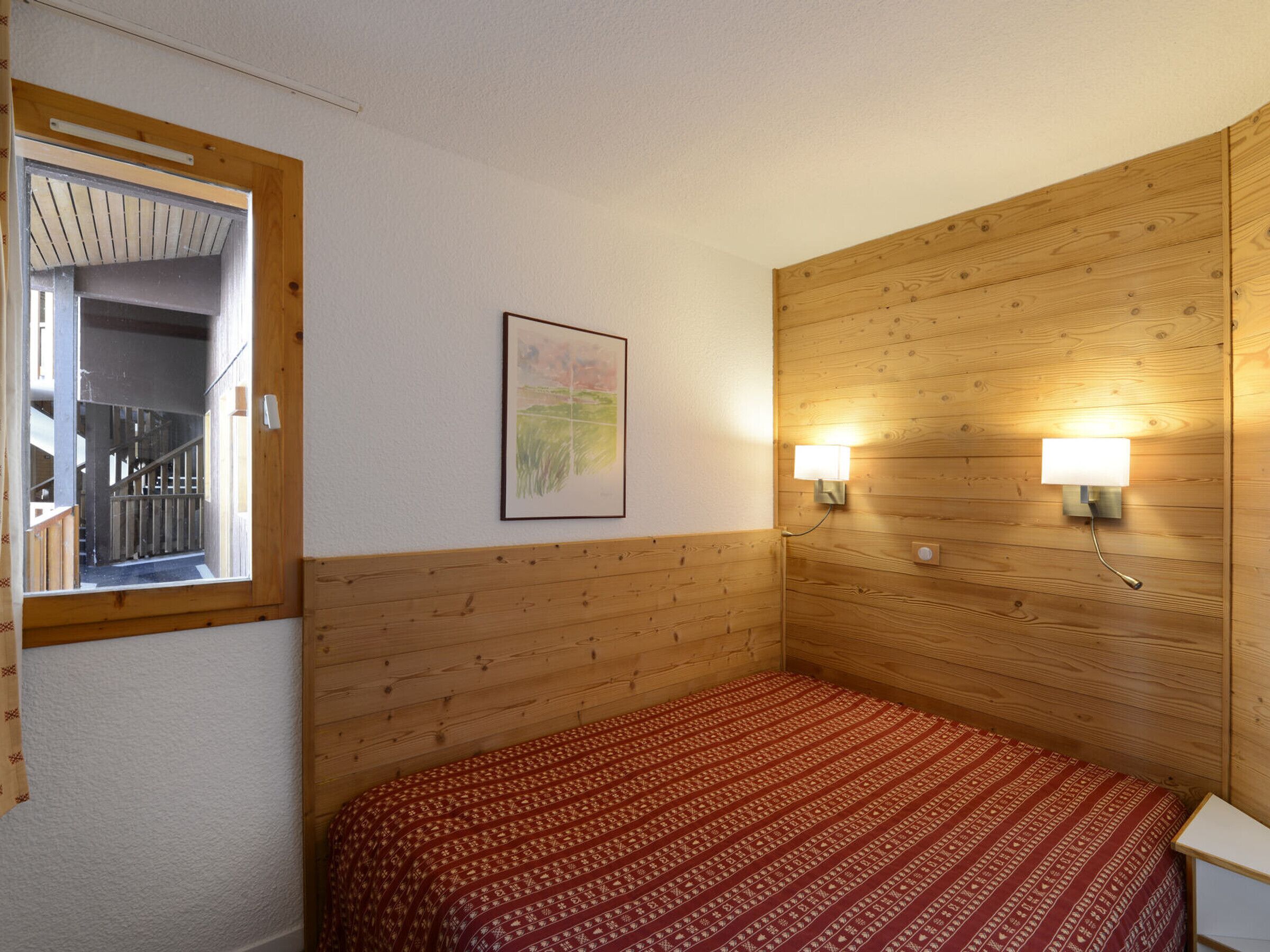 Een slaapkamer met een houten hoofdbord, een bed met een geborduurde spreideken, een ingelijst schilderij aan de muur en twee wandlampen.