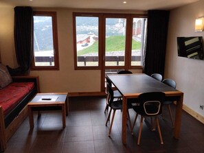 A room with a bed, a small table, a dining table with chairs, and large windows with a view of a village and mountains. - Renovated ski-in apartment with free parking, pets allowed (La Plagne Montchavin Les Coches)