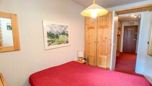 A bedroom with a red bedspread, a wooden wardrobe, a bedside lamp, and a framed landscape painting on the wall.