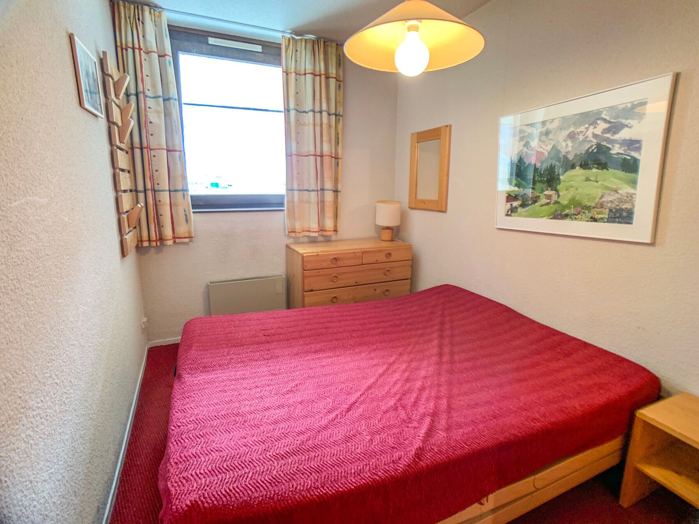 A bedroom with a bed, a dresser, a lamp, a window with curtains, and a framed picture on the wall.