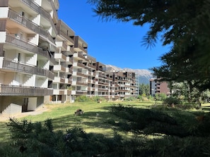 Un edificio residencial de varios pisos con balcones, un césped cuidado y montañas al fondo.