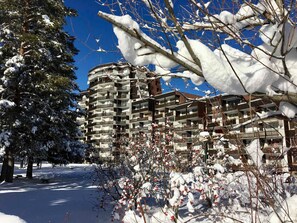 A multi-story hotel with balconies, surrounded by snow-covered trees and a clear blue sky.