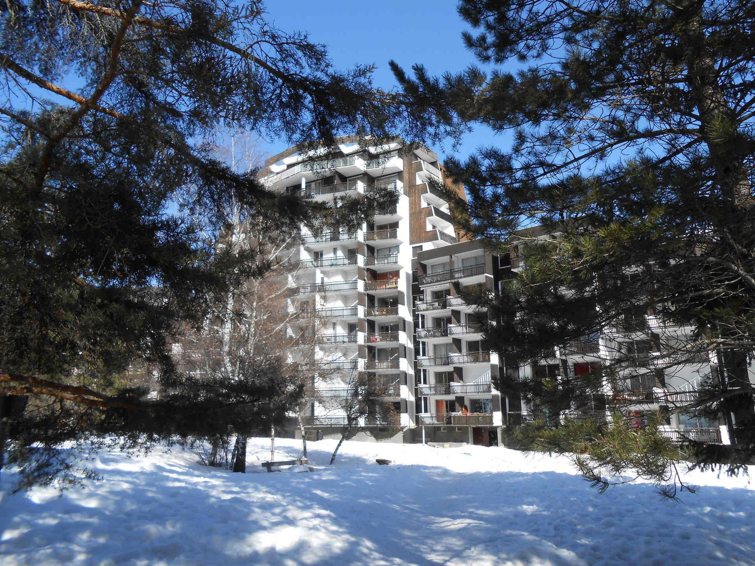 A multi-story residential building with balconies, surrounded by snow and pine trees.