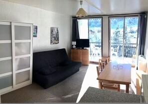 A modern living room with a black sofa, a wooden dining table, and a TV on a wooden cabinet. Large windows offer a view of a snowy landscape.