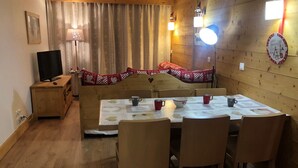 Une pièce chaleureuse avec une table à manger en bois, préparée pour quatre personnes, un canapé agrémenté de coussins à motifs de cœurs, un téléviseur posé sur un meuble en bois et une horloge murale.