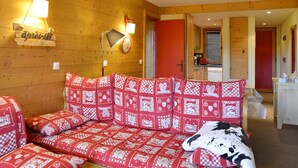 Une pièce chaleureuse avec un mur en bois, un canapé orné de coussins à carreaux, une lampe fixée au mur et une porte rouge donnant sur une cuisine.