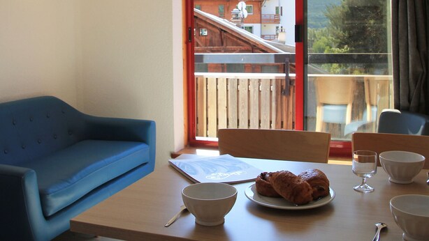 A room with a blue sofa, a wooden table with a plate of croissants, bowls, and a glass. A window reveals a mountain view. - Bright apartment near ski lifts, mountain corner, pets allowed (Briançon)