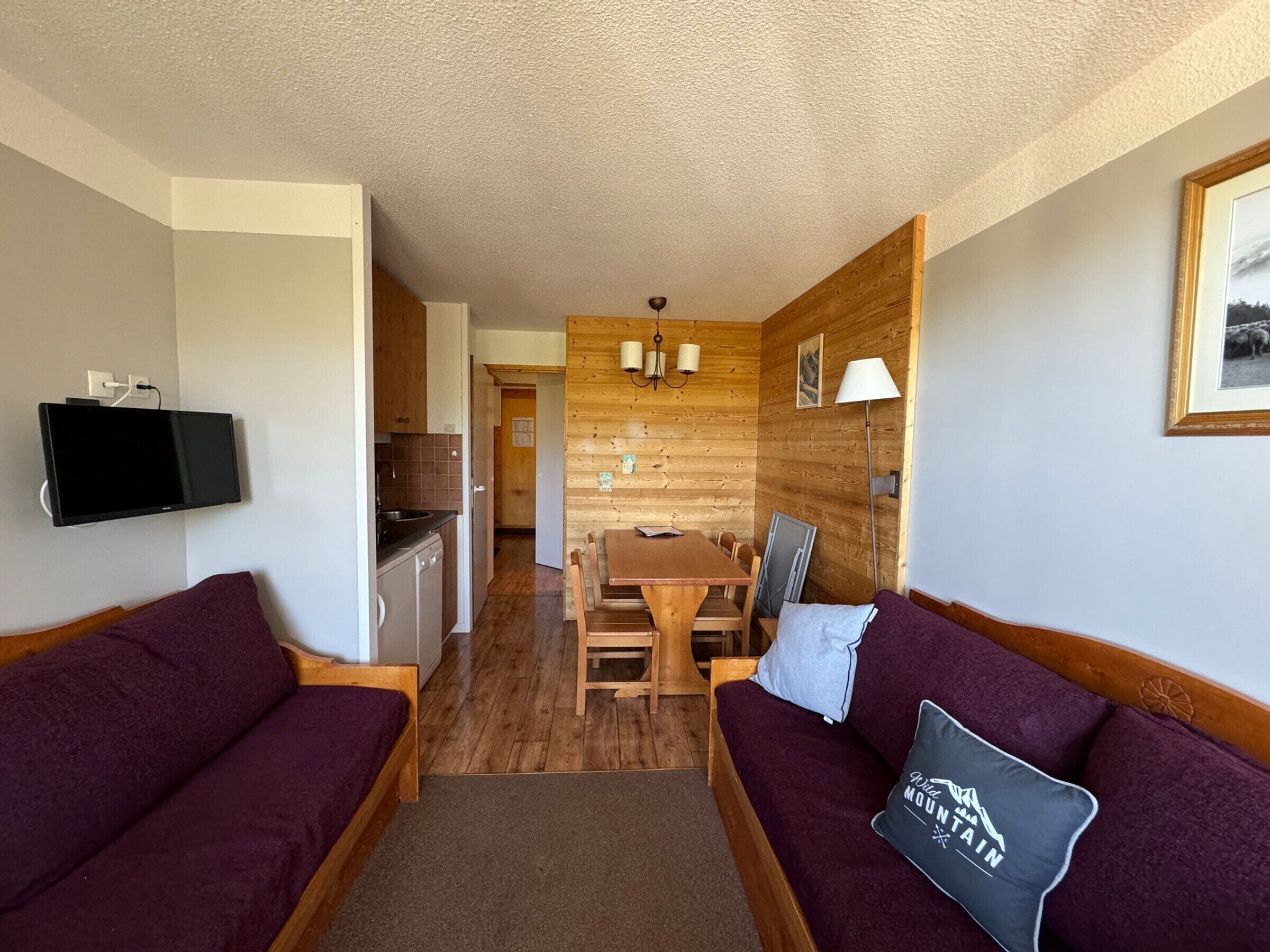 Una habitación compacta con una mesa de comedor de madera, dos sofás de madera, un televisor montado en la pared y una lámpara.