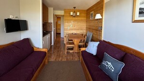A compact room with a wooden dining table, two wooden sofas, a TV mounted on the wall, and a lamp.