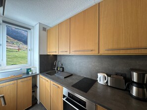 A modern kitchen with wooden cabinets, a built-in oven, a coffee maker, and a view of a green landscape through a window.