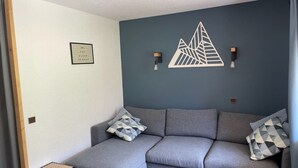 A modern living room with a grey sofa, patterned cushions, a small side table, and a wall-mounted light fixture. - Renovated Studio 2019 · Ski-in Ski-out · Balcony (Belle Plagne)