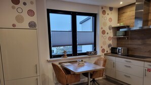 A compact kitchen with a dining table, chairs, and a window with blinds. - Vacation home with garden and terrace, 15 minutes from the Moselle (Blankenrath)