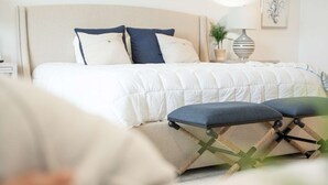 En pænt redt seng med hvid dyne og to puder, et sengebord med lampe og en lille plante på skuffesektionen.