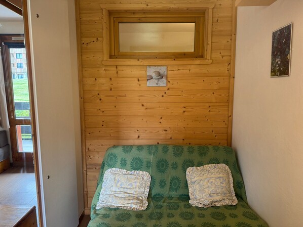 Ein Zimmer mit einer Holzwand, einem Fenster, einem grünen Sofa mit gemusterten Kissen und einem kleinen Bilderrahmen.
