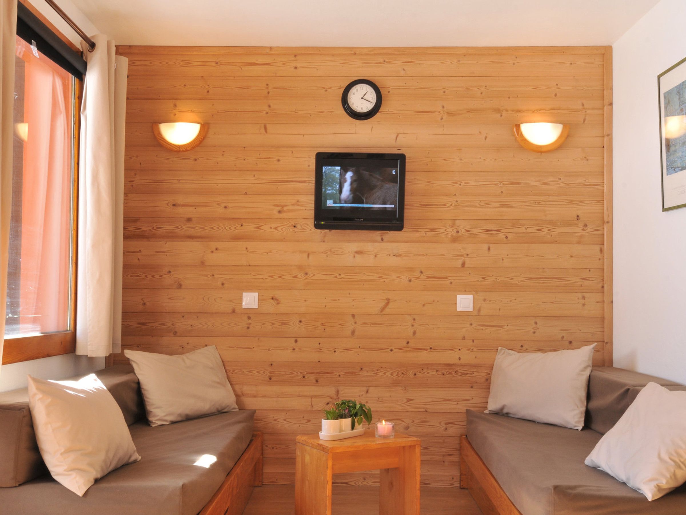 Ein gemütliches Zimmer mit Holzwandverkleidung, einem an der Wand montierten Fernseher, zwei Sofas mit Kissen, einem kleinen Holztisch mit Kerze und Pflanze sowie einer Uhr über dem Fernseher.