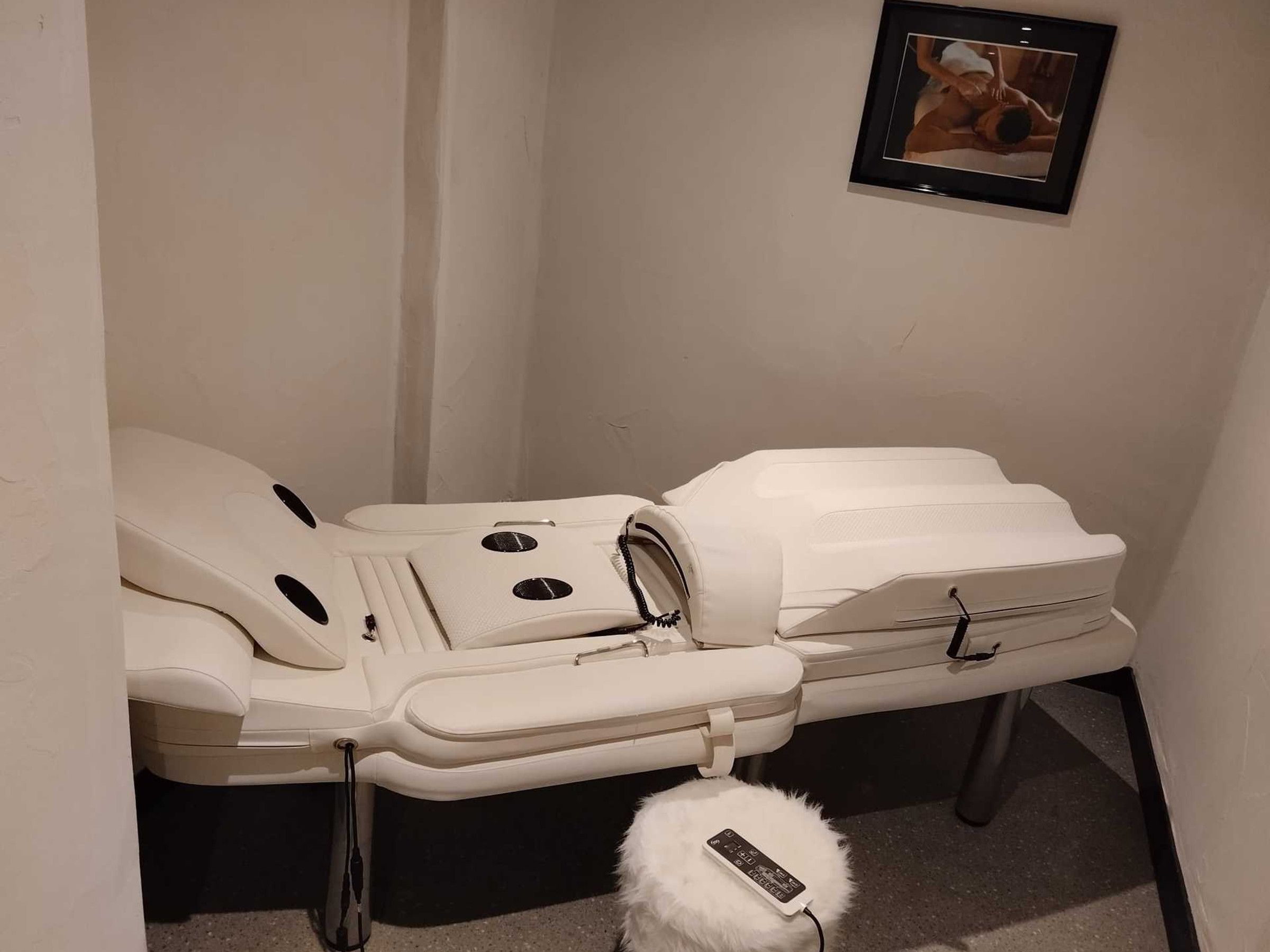 Ett rum med en massagebänk, en liten, mjuk pall och en inramad tavla på väggen.
