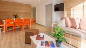 Un salon moderne avec un mur en bois, un canapé avec des coussins, une table basse avec un magazine et une plante, et un coin repas avec des chaises et une table orange.