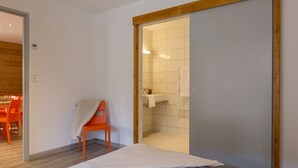 Une chambre à coucher avec un lit, une porte coulissante à cadre en bois, une petite chaise orange et une salle de bain visible, dotée d’un sol carrelé et d’un porte-serviettes.