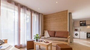Un salon moderne comprenant un canapé, une table basse et un pouf, un petit coin cuisine avec des appareils électroménagers, et de grandes fenêtres ornées de rideaux légers.