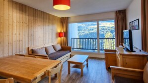 Habitación con piso de madera, un sofá, una mesa de comedor, un televisor y un balcón con vistas al bosque.