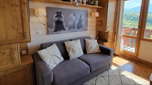 Ein gemütliches Wohnzimmer mit grauem Sofa, Holzschränken und einem großen Fenster mit Blick ins Freie.