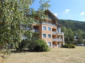A multi-story building with balconies, surrounded by trees and a hilly landscape.