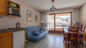 A compact living space with a kitchenette, a blue sofa, a dining table with chairs, and a view through a glass door. - Cozy studio with ski shuttle and private locker - 4 guests (La Salle-les-Alpes)