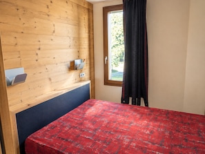A room with a wooden wall, a window with a view of greenery, a bed with a red patterned bedspread, and a small shelf with a cup and a book. - Renovated Apartment 5 ppl - Pets Allowed, Free Parking (La Plagne Montchavin Les Coches)